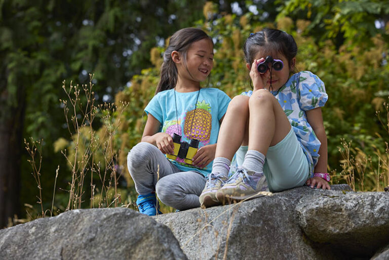 Kids enjoying nature at summer camp while using binoculars, sitting on rocks amidst trees. Kids enjoying nature at summer camp while using binoculars, sitting on rocks amidst trees.