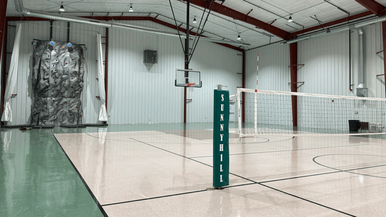 Indoor multisport court with basketball hoop, volleyball net, and rock wall at summer camp facility. Indoor multisport court with basketball hoop, volleyball net, and rock wall at summer camp facility.