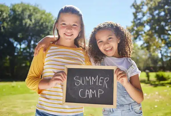 Two smiling girls holding a Summer Camp sign outdoors on a sunny day, promoting camp activities.