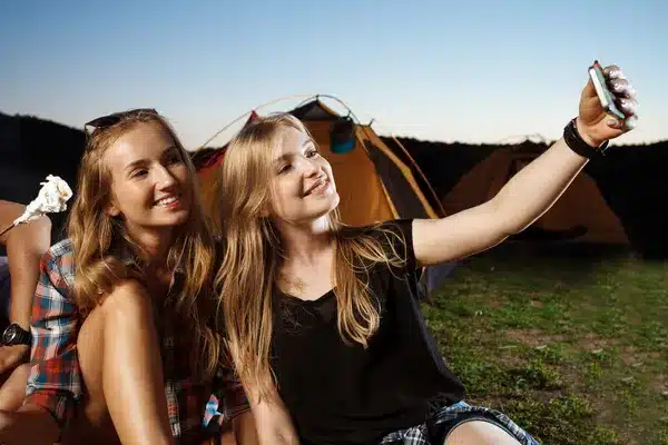 Teen girls taking a selfie at summer camp, with tents in the background at sunset. Perfect summer adventure experience.