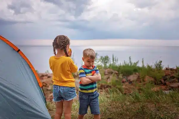 Children camping by a lake, enjoying nature at summer camp. Tent set up on grassy shore, scenic view in background.