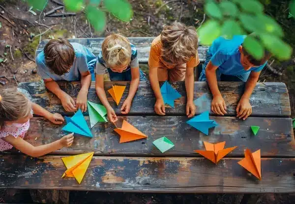 Children making paper airplanes at summer camp, enjoying outdoor crafting activities.
