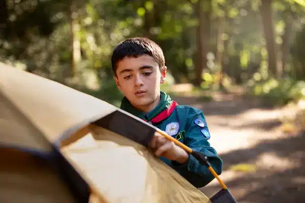 Young camper setting up a tent in a forested summer camp setting.