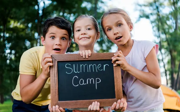 Three kids holding a chalkboard with Summer Camp written on it, surrounded by trees. Fun outdoor activities await!