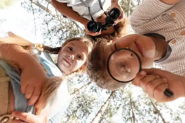 Kids exploring nature with magnifying glass and binoculars at summer camp.