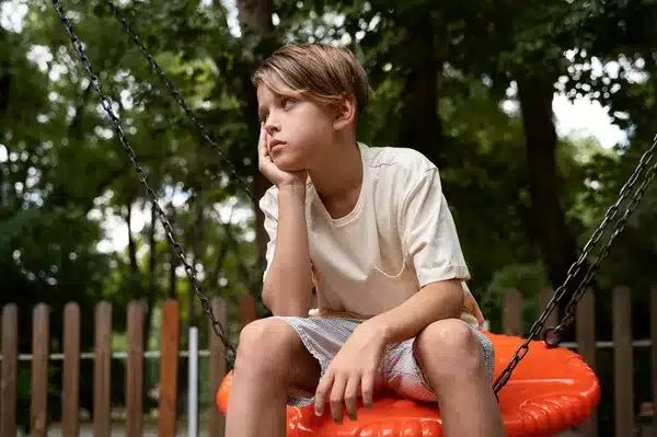 Child sitting on a swing at summer camp playground, daydreaming in a peaceful setting with trees in the background.