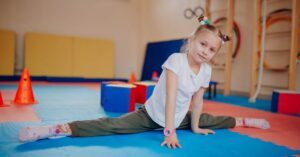 Child practicing gymnastics splits at summer camp in indoor play area.