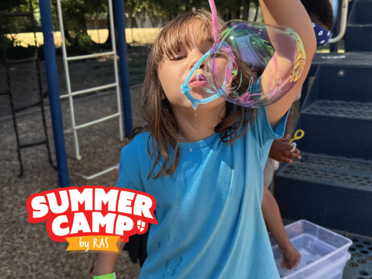 Child at summer camp blowing large soap bubble with wand on playground, colorful bubble and blue camp shirt Child at summer camp blowing large soap bubble with wand on playground, colorful bubble and blue camp shirt