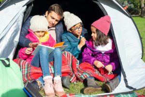 Kids and counselor reading in a tent at summer camp, bundled in jackets and beanies on a plaid blanket